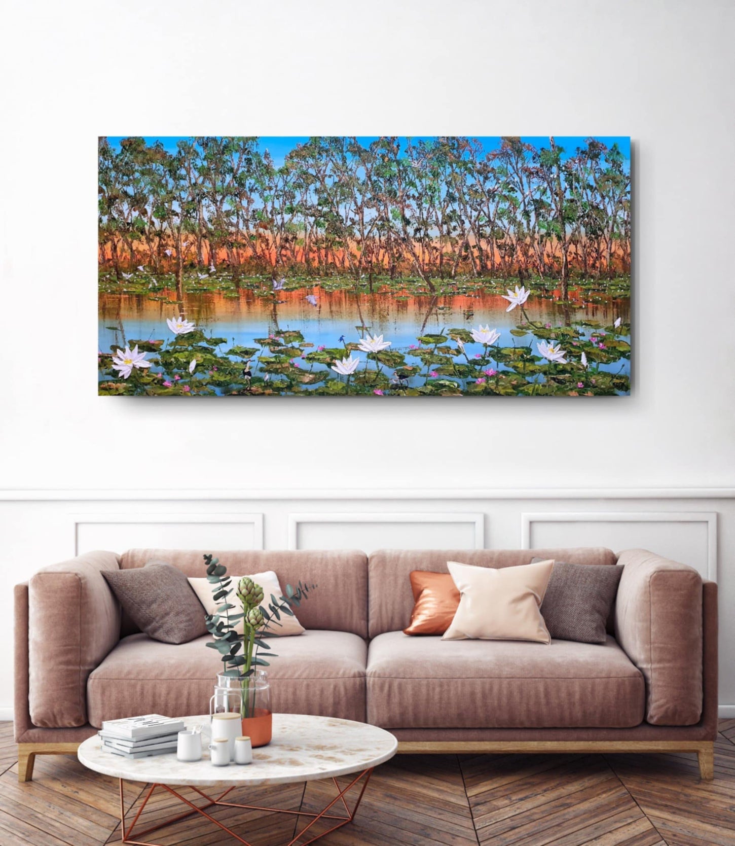 Panoramic landscape painting of a lake with trees and flowers above a beige sofa.
